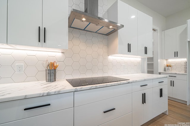 kitchen featuring backsplash, black electric stovetop, white cabinets, wall chimney exhaust hood, and light stone counters