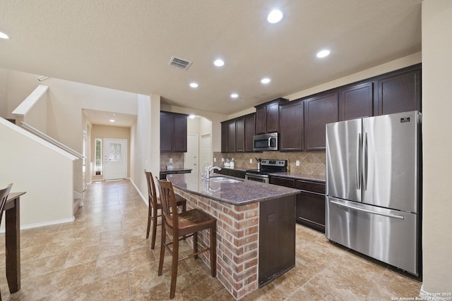 kitchen featuring sink, dark stone countertops, an island with sink, appliances with stainless steel finishes, and dark brown cabinets