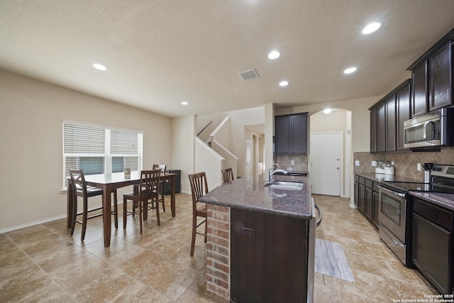 kitchen featuring backsplash, sink, an island with sink, appliances with stainless steel finishes, and dark brown cabinets