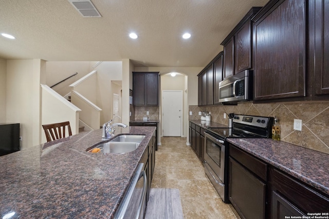 kitchen with appliances with stainless steel finishes, backsplash, dark stone counters, dark brown cabinetry, and sink