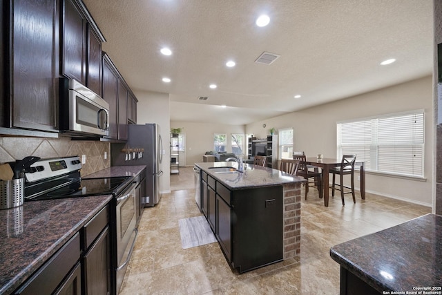 kitchen featuring backsplash, dark stone counters, stainless steel appliances, sink, and an island with sink