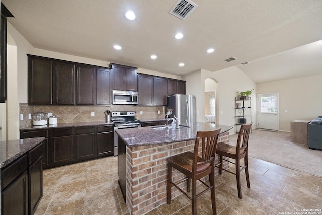 kitchen with dark brown cabinetry, stainless steel appliances, dark stone countertops, decorative backsplash, and a center island with sink