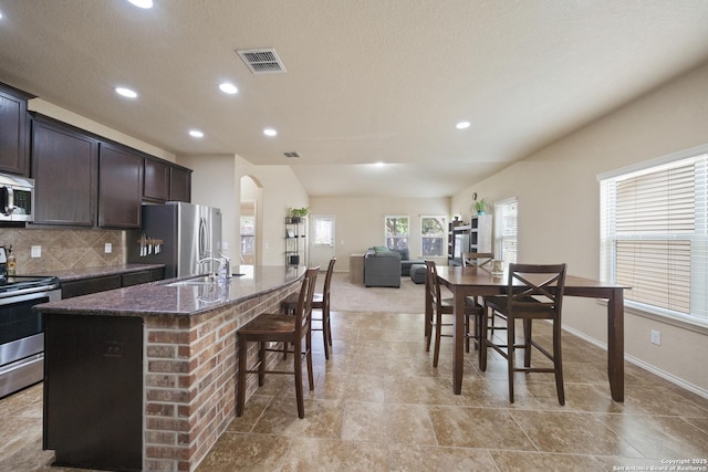 kitchen with decorative backsplash, appliances with stainless steel finishes, dark stone counters, dark brown cabinetry, and a kitchen island with sink