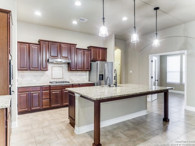 kitchen featuring light stone counters, an island with sink, stainless steel appliances, and decorative light fixtures