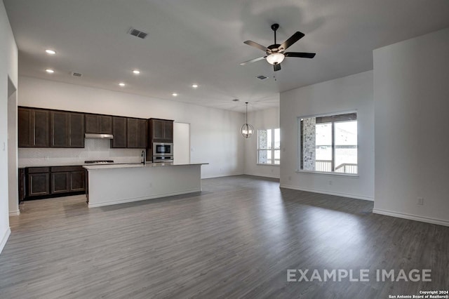 kitchen featuring stainless steel microwave, backsplash, a kitchen island with sink, hanging light fixtures, and hardwood / wood-style flooring