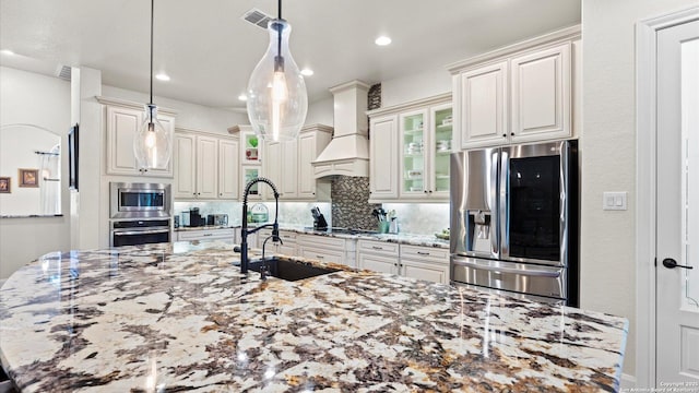 kitchen featuring light stone countertops, sink, stainless steel appliances, premium range hood, and decorative light fixtures