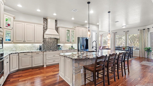 kitchen with light stone countertops, custom exhaust hood, pendant lighting, a center island with sink, and stainless steel fridge with ice dispenser