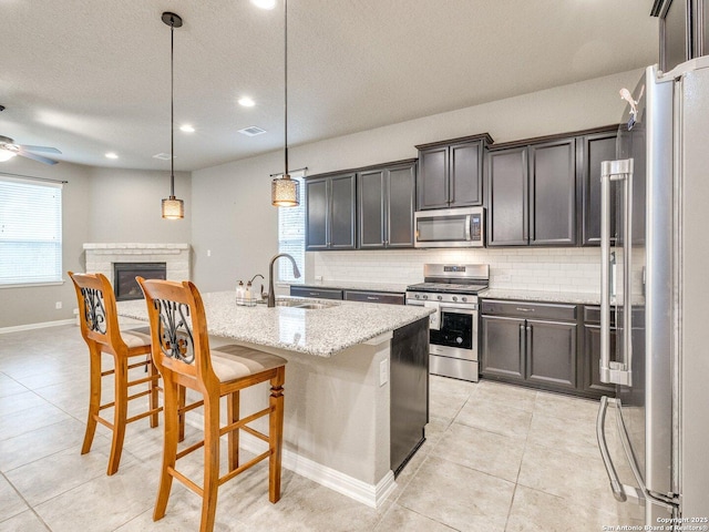 kitchen with light stone counters, stainless steel appliances, sink, pendant lighting, and an island with sink