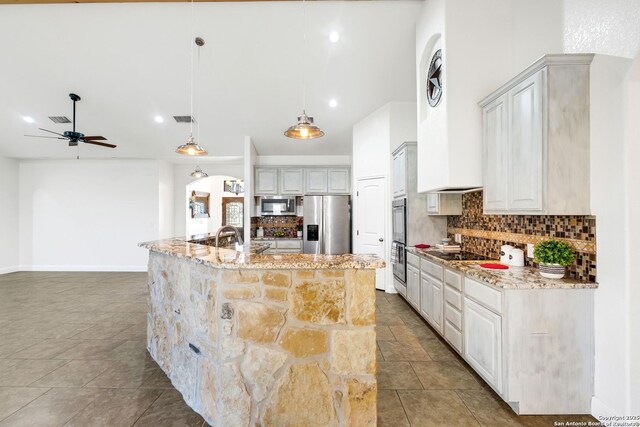 kitchen with decorative backsplash, appliances with stainless steel finishes, light stone countertops, decorative light fixtures, and a center island with sink