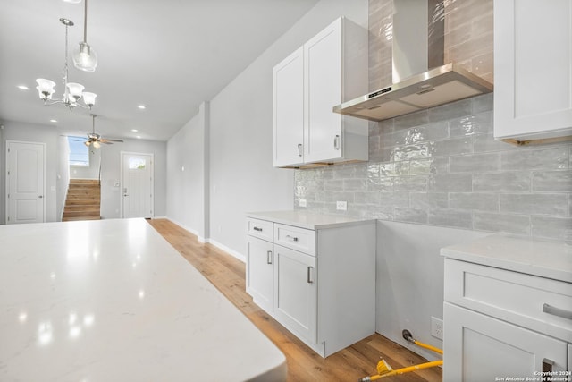 kitchen featuring light stone counters, ceiling fan with notable chandelier, wall chimney range hood, pendant lighting, and white cabinets