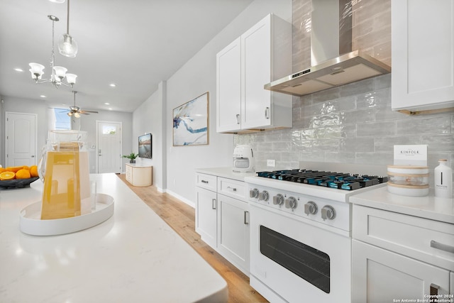 kitchen featuring white gas range oven, tasteful backsplash, ceiling fan with notable chandelier, wall chimney range hood, and white cabinetry