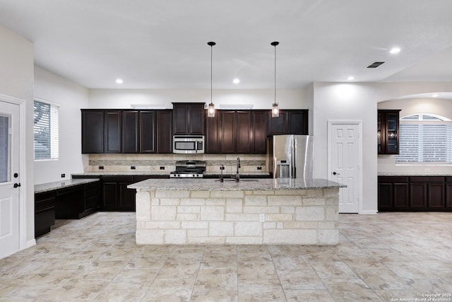 kitchen featuring light stone counters, dark brown cabinetry, stainless steel appliances, pendant lighting, and an island with sink