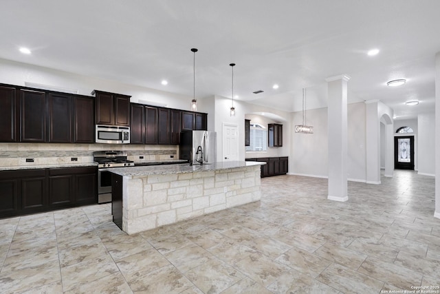 kitchen featuring appliances with stainless steel finishes, tasteful backsplash, light stone counters, decorative light fixtures, and an island with sink