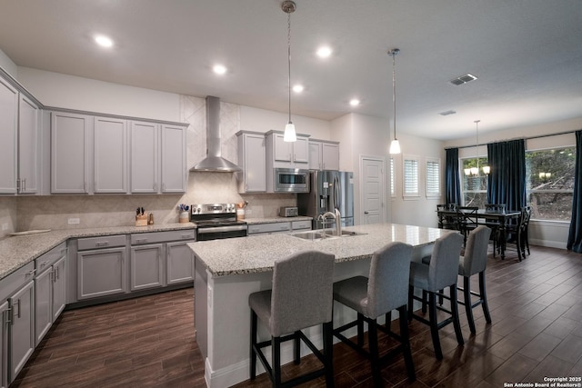 kitchen with wall chimney range hood, light stone counters, pendant lighting, a center island with sink, and appliances with stainless steel finishes