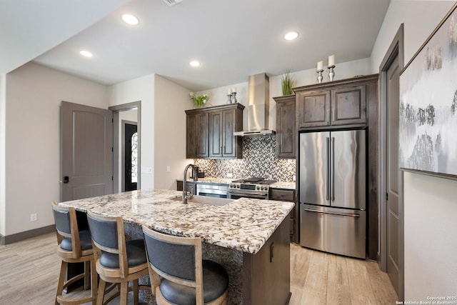 kitchen with appliances with stainless steel finishes, light hardwood / wood-style floors, a kitchen island with sink, and wall chimney range hood