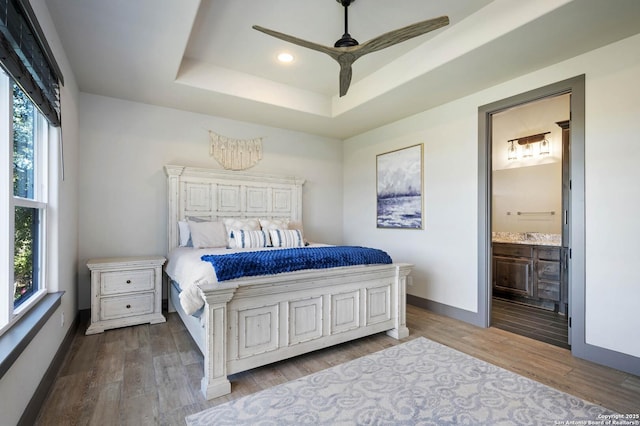 bedroom featuring ceiling fan, a raised ceiling, ensuite bathroom, hardwood / wood-style floors, and multiple windows