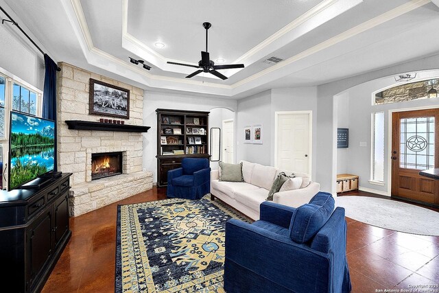 living room featuring a tray ceiling, a stone fireplace, ceiling fan, and crown molding