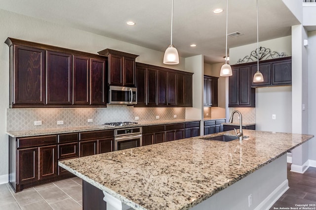 kitchen with a center island with sink, sink, hanging light fixtures, light stone countertops, and stainless steel appliances