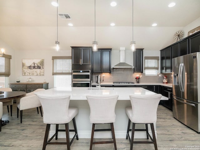 kitchen with pendant lighting, stainless steel appliances, a center island with sink, and wall chimney range hood