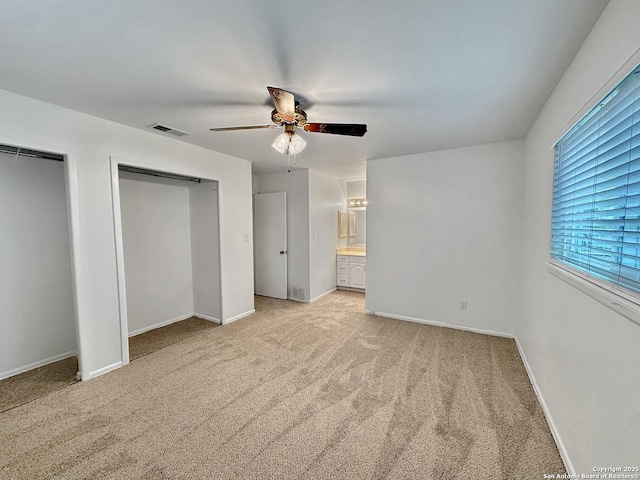 unfurnished bedroom with ceiling fan, light colored carpet, ensuite bathroom, and two closets