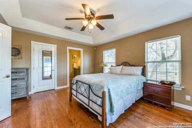 bedroom with hardwood / wood-style flooring, ceiling fan, ensuite bathroom, and a tray ceiling