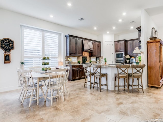 kitchen featuring decorative backsplash, an island with sink, built in microwave, dark brown cabinets, and a breakfast bar area