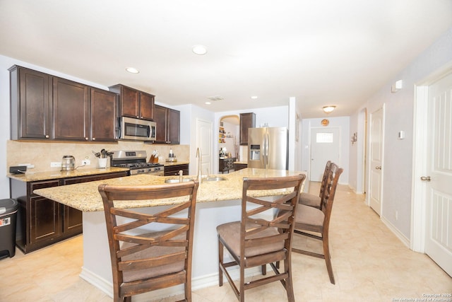 kitchen featuring a kitchen island with sink, sink, appliances with stainless steel finishes, dark brown cabinets, and light stone counters