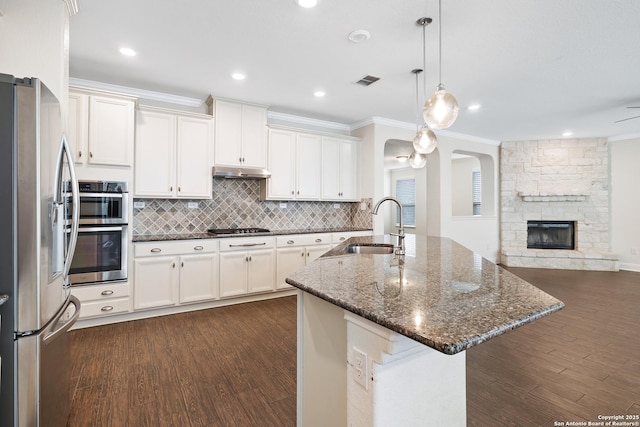 kitchen featuring appliances with stainless steel finishes, a center island with sink, dark stone countertops, a fireplace, and hanging light fixtures