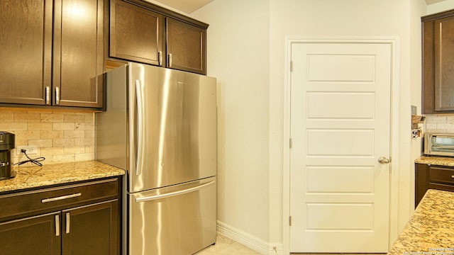 kitchen featuring decorative backsplash, dark brown cabinetry, light stone countertops, and stainless steel refrigerator