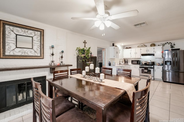 tiled dining space featuring a brick fireplace, ceiling fan, crown molding, and sink