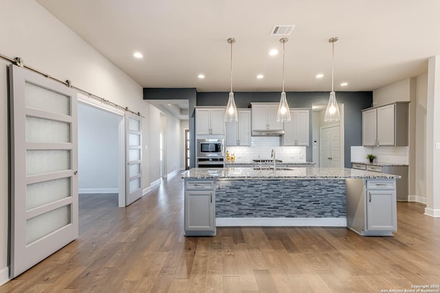 kitchen featuring light stone countertops, appliances with stainless steel finishes, a barn door, a center island with sink, and decorative light fixtures