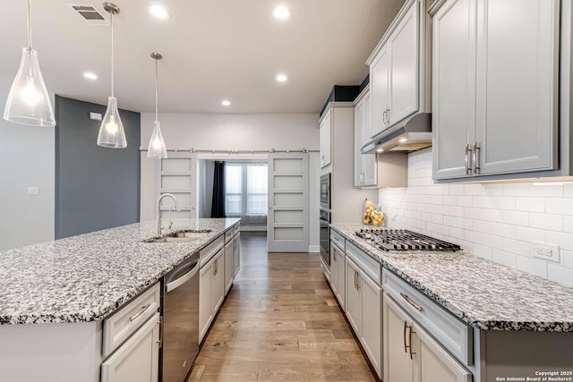 kitchen with sink, stainless steel appliances, hanging light fixtures, light stone counters, and a kitchen island with sink