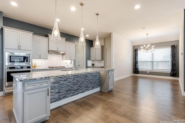 kitchen featuring a kitchen island with sink, hanging light fixtures, sink, light stone counters, and stainless steel appliances