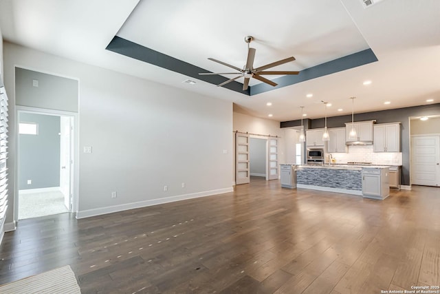 unfurnished living room with a tray ceiling, a barn door, ceiling fan, and dark hardwood / wood-style flooring