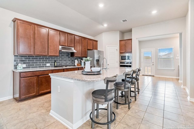 kitchen with a center island with sink, decorative backsplash, light stone counters, appliances with stainless steel finishes, and a kitchen bar