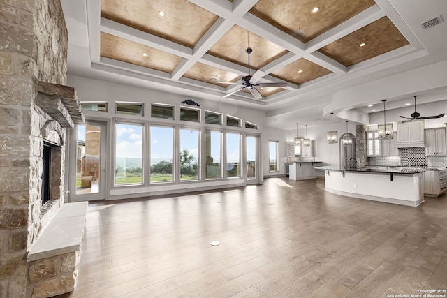 unfurnished living room featuring coffered ceiling, ceiling fan with notable chandelier, a stone fireplace, light hardwood / wood-style flooring, and beam ceiling