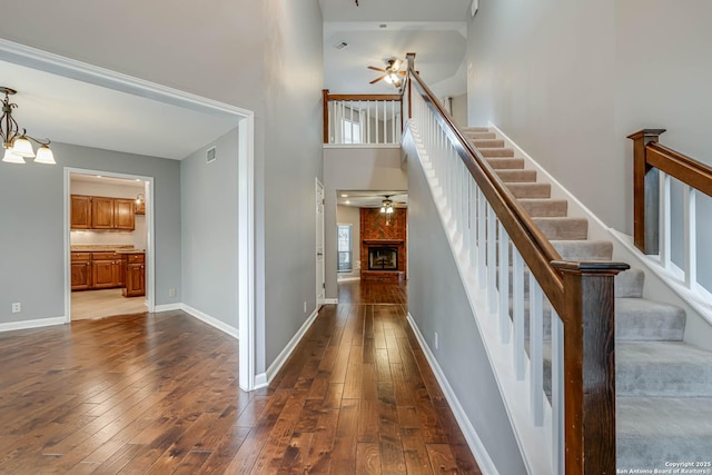 staircase with a fireplace, a towering ceiling, ceiling fan with notable chandelier, and hardwood / wood-style flooring