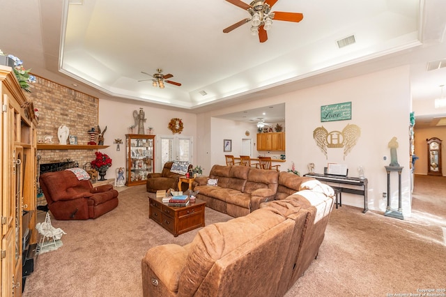 carpeted living room with a brick fireplace, ceiling fan, and a tray ceiling