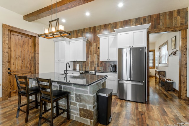 kitchen with beamed ceiling, dark stone countertops, stainless steel fridge, a center island with sink, and white cabinets