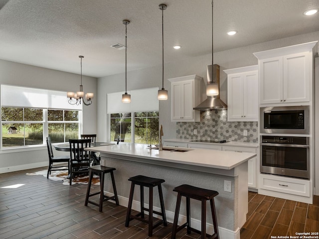 kitchen featuring wall chimney exhaust hood, stainless steel oven, a kitchen island with sink, built in microwave, and hanging light fixtures