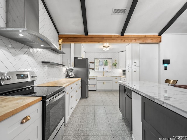 kitchen featuring beam ceiling, butcher block counters, wall chimney exhaust hood, stainless steel appliances, and white cabinets