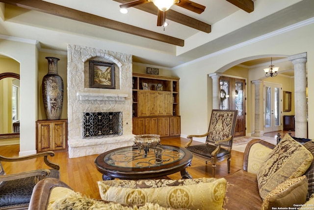 living room with light wood-type flooring, ornate columns, ornamental molding, ceiling fan with notable chandelier, and a stone fireplace