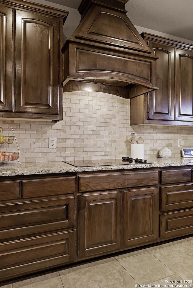 kitchen with black electric stovetop, tasteful backsplash, light stone counters, custom range hood, and dark brown cabinets