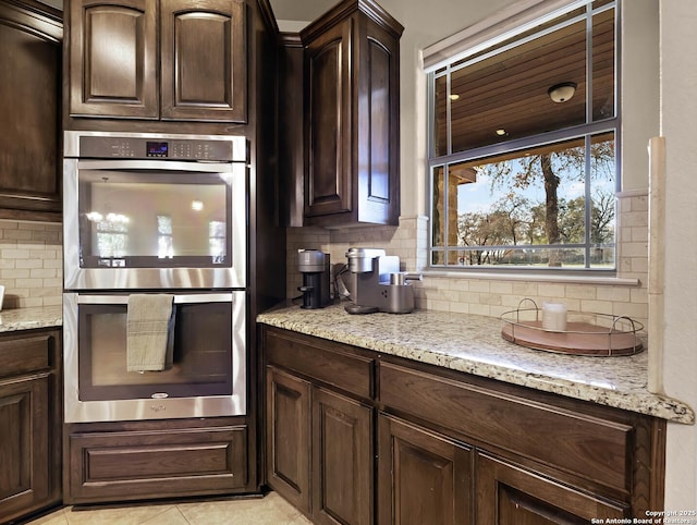 kitchen featuring double oven, backsplash, light stone countertops, and dark brown cabinetry