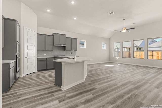 kitchen featuring gray cabinets, backsplash, a center island with sink, vaulted ceiling, and stainless steel oven