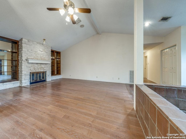 unfurnished living room featuring hardwood / wood-style flooring, ceiling fan, vaulted ceiling with beams, a textured ceiling, and a stone fireplace