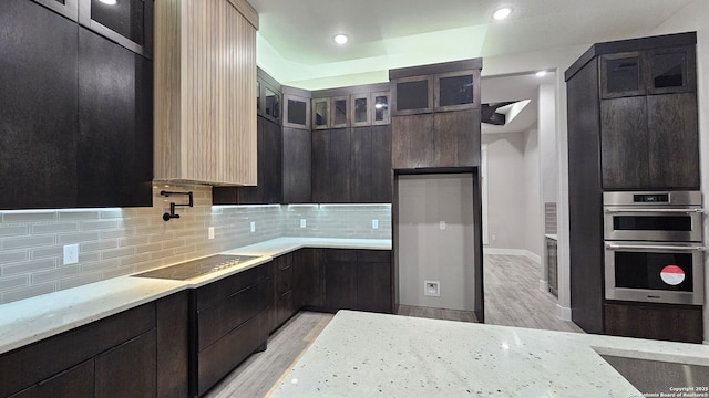 kitchen with black electric stovetop, double oven, backsplash, light stone counters, and light hardwood / wood-style flooring