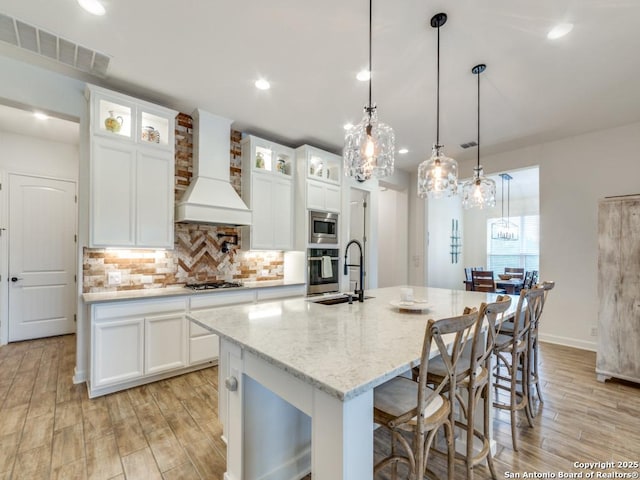 kitchen featuring a center island with sink, white cabinets, premium range hood, and stainless steel appliances