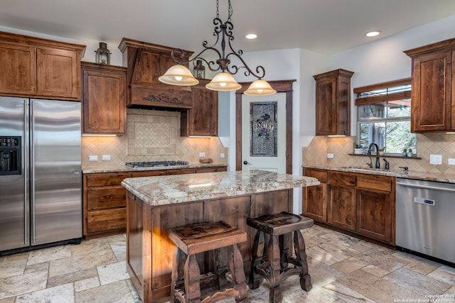 kitchen with sink, stainless steel appliances, light stone counters, decorative light fixtures, and a kitchen island