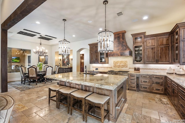 kitchen with custom range hood, a kitchen island with sink, glass insert cabinets, and stone tile flooring